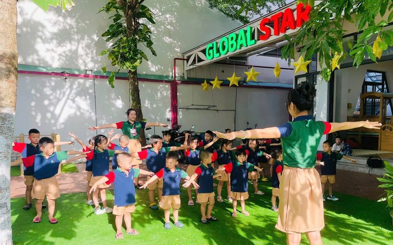Các bé dang tay tập thể dục theo cô trên sân cỏ trước cổng trường