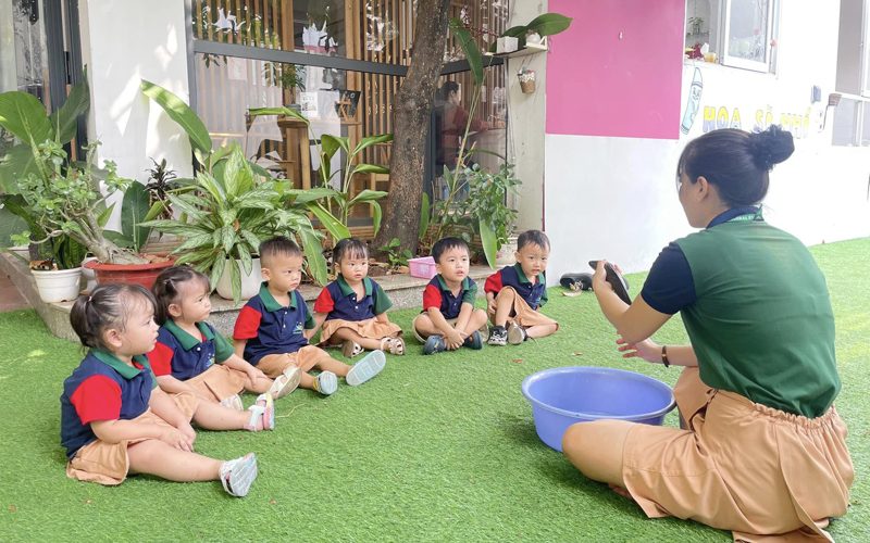 Các bé ngồi trên thảm cỏ ngoài sân nghe cô hướng dẫn trò chơi