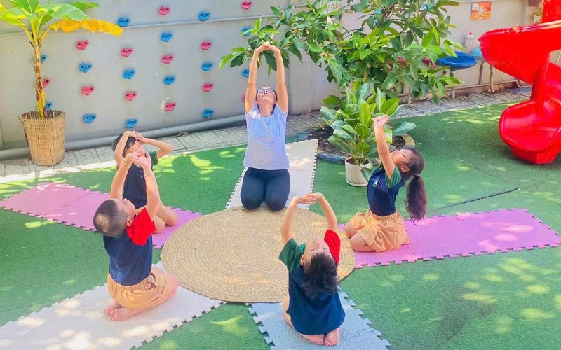 Cô và các bé giơ tay tập vận động trên thảm ở sân chơi